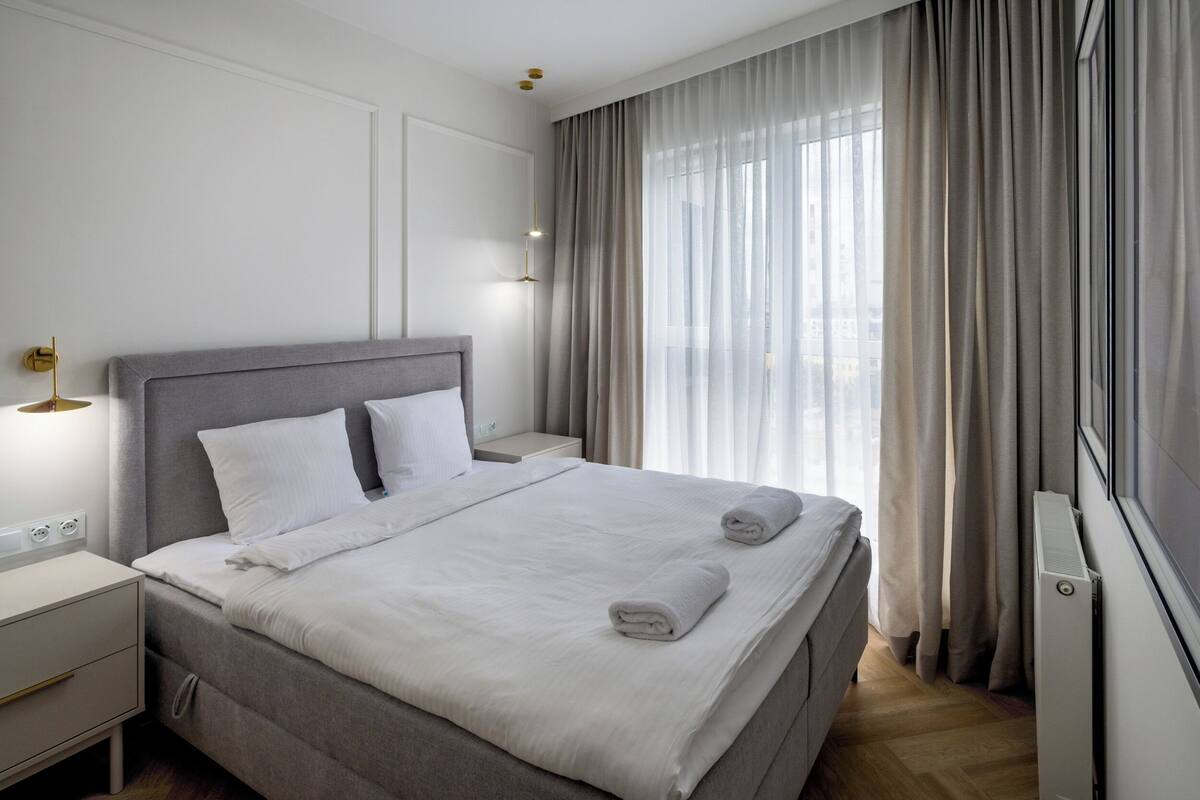 A modern hotel room with a large bed, bedside tables, and a view of the city through sheer curtains.