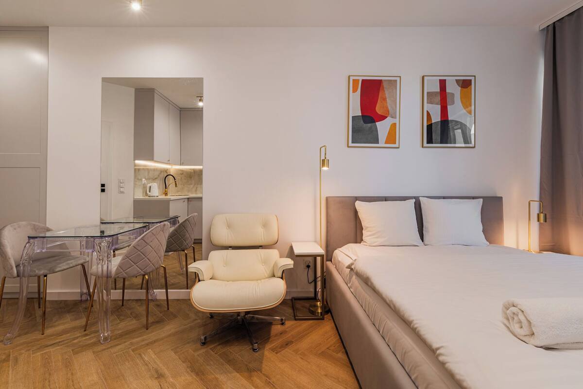 A hotel room with a bed, a desk with a chair, a small table, and a chair. There are two paintings on the wall.