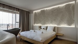 A modern hotel room with a large bed, a wooden floor, a view of the city, a bathtub, and a large textured wall. - RentPlanet - Apartamenty Quorum (Wroclaw)