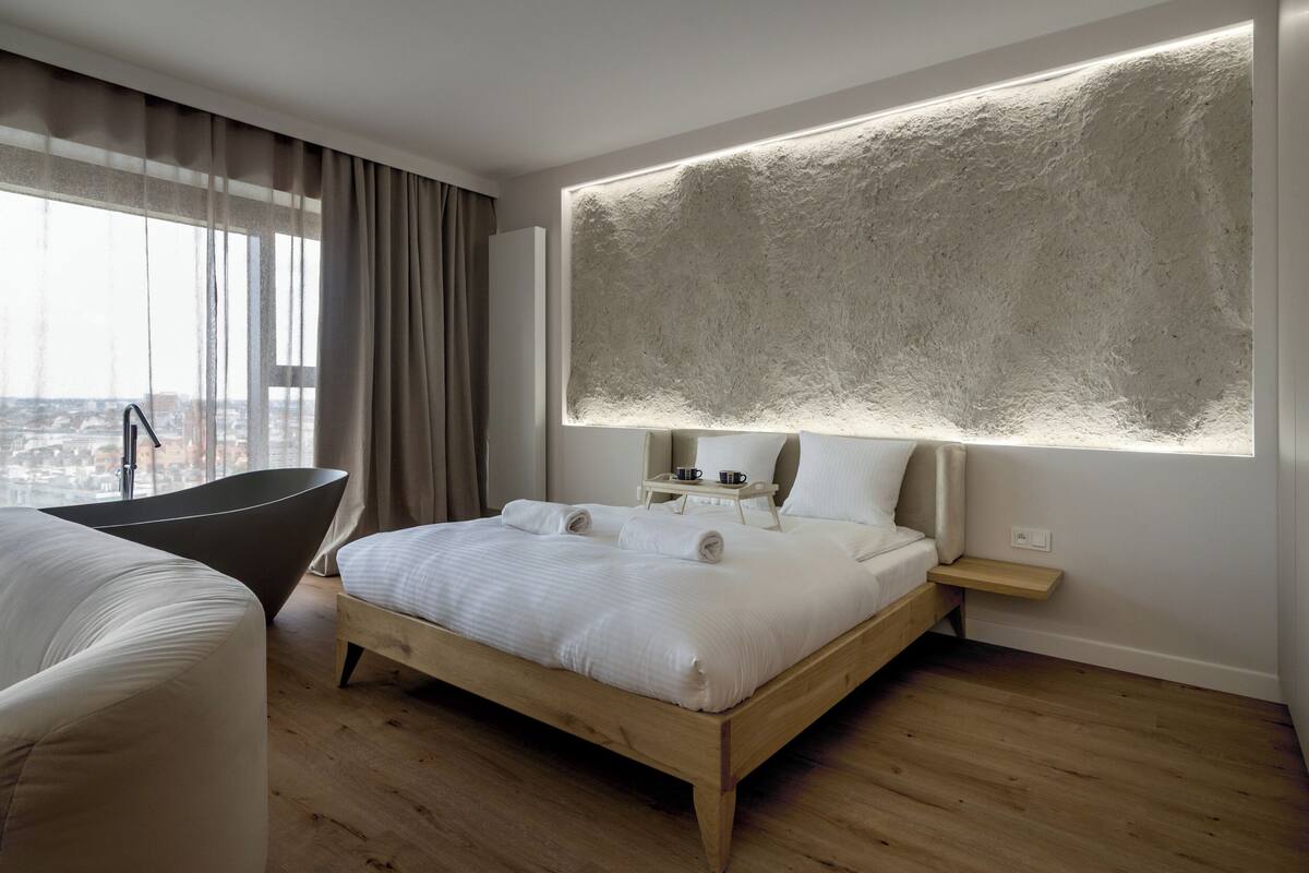 A modern hotel room with a large bed, a wooden floor, a view of the city, a bathtub, and a large textured wall.