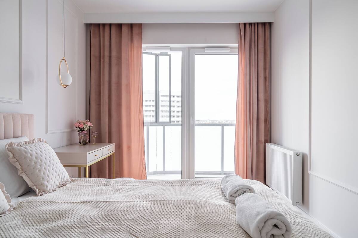 A bedroom with a bed, a bedside table with a vase of flowers, a radiator, and a window with curtains.