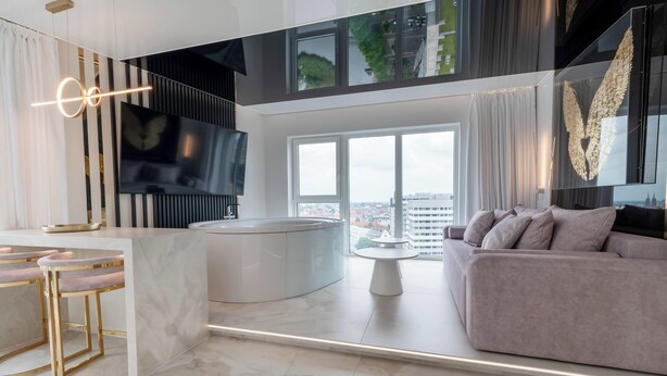 A modern living room with a large window, a sofa, a TV, and a kitchen area with bar stools. - RentPlanet - Apartamenty Quorum (Wroclaw)