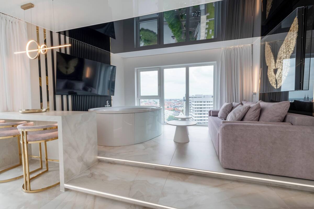 A modern living room with a large window, a sofa, a TV, and a kitchen area with bar stools.