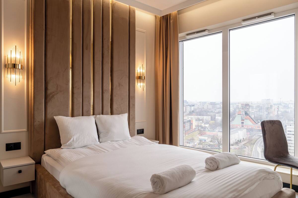 A hotel room with a large bed, two bedside tables, a chair, and a view of the city through large windows.