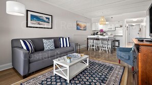 Ein modernes Wohnzimmer mit grauem Sofa, weißem Couchtisch und einem gemusterten Teppich. Im Hintergrund ist eine Küche mit weißen Schränken und ein Essbereich mit weißen Stühlen erkennbar.