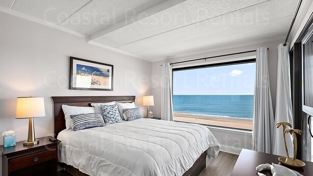 Ein Hotelzimmer mit einem großen Bett, zwei Nachttischen, einem Wandbild in einem Rahmen und Blick auf den Ozean durch ein großes Fenster.
