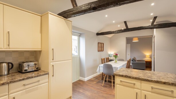A kitchen with white cabinets, a granite countertop, and a toaster. There is a dining area with a table and chairs, and a window with blinds. - Host Stay Croft Cottage (Malton)