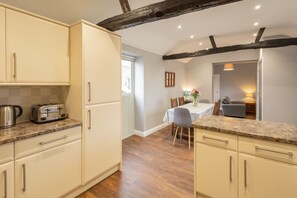 A kitchen with white cabinets, a granite countertop, and a toaster. There is a dining area with a table and chairs, and a window with blinds. - Host Stay Croft Cottage (Malton)