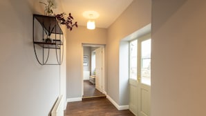 A hallway with wooden flooring, a radiator, a door with a window, and a wall-mounted shelf with a plant. - Host Stay Croft Cottage (Malton)