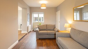 A modern living room with a grey sofa, a small table with a lamp, and a large mirror on the wall. - Host Stay Croft Cottage (Malton)