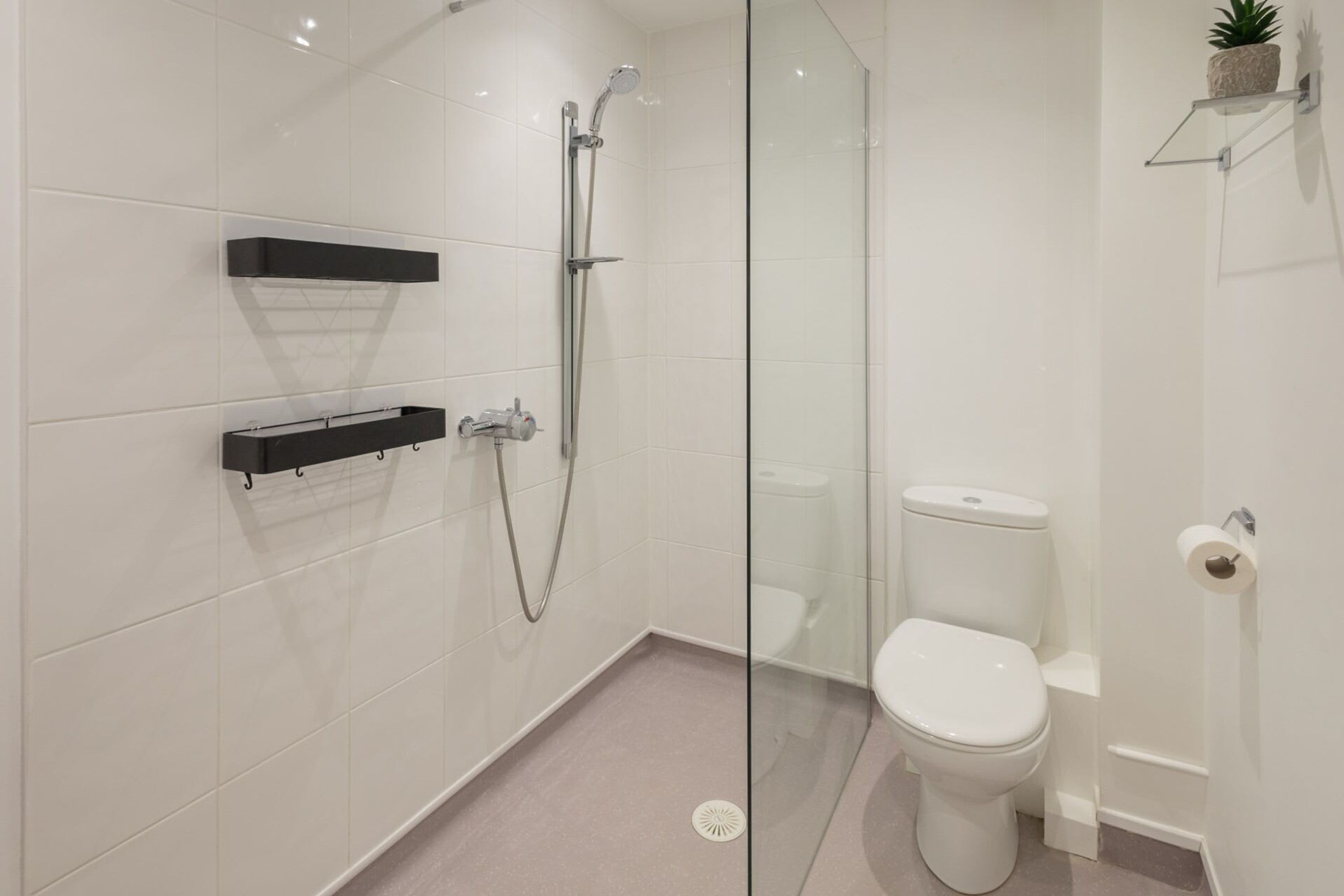 A modern bathroom with a glass shower enclosure, a toilet, and two black floating shelves.