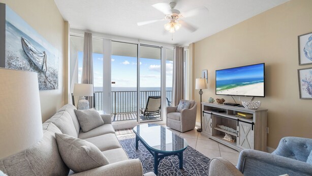 A spacious living room with a large sofa, a coffee table, and a television. There is a balcony with a view of the ocean.