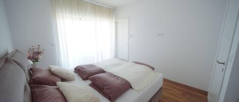 Un dormitori amb un llit, almoines, una taula de nit amb un vas de flors i una finestra amb cortines.
