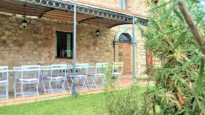 A stone building with a covered patio area, outdoor seating with white chairs, and a wooden door. - Campo Al Vento (Monte Castello di Vibio)