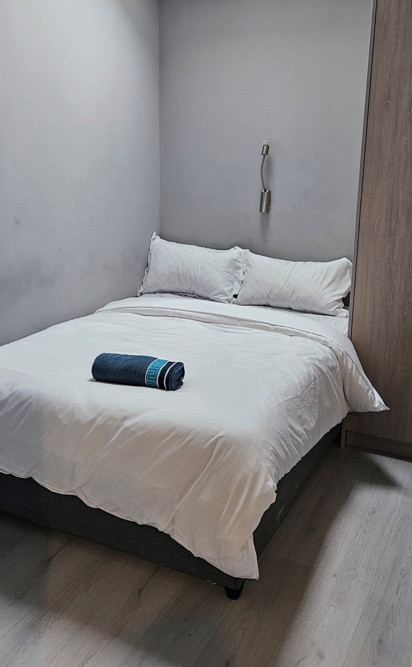 Un letto singolo con materasso e cuscini bianchi, un asciugamano blu arrotolato e un lampadario a parete.