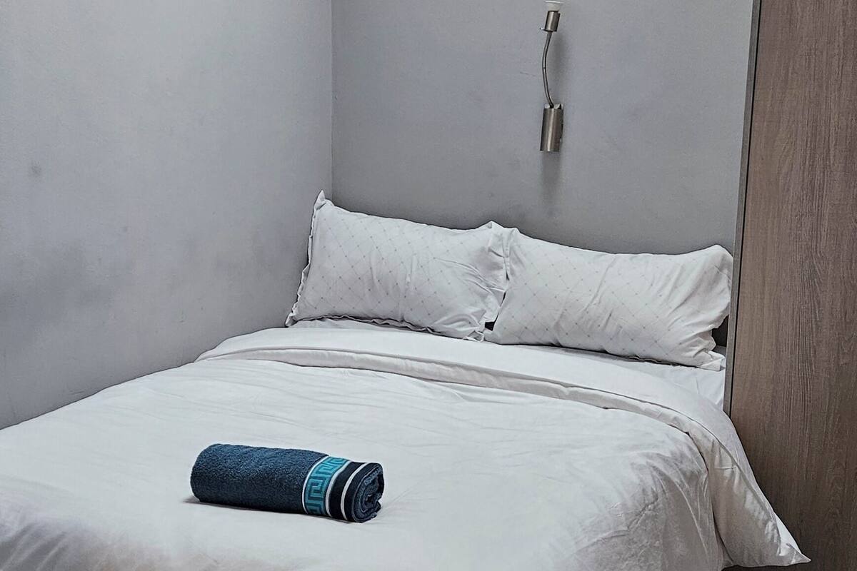 A single bed with a white mattress and pillows, a rolled blue towel, and a wall-mounted light fixture.