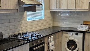 A kitchen with light wood cabinets, a gas stove, an oven, a washing machine, and a window with blinds.