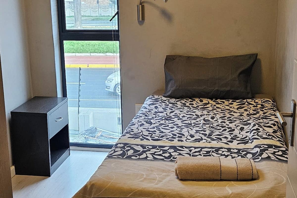 A bedroom with a bed, a nightstand, a window with a view, and a wall-mounted light fixture.