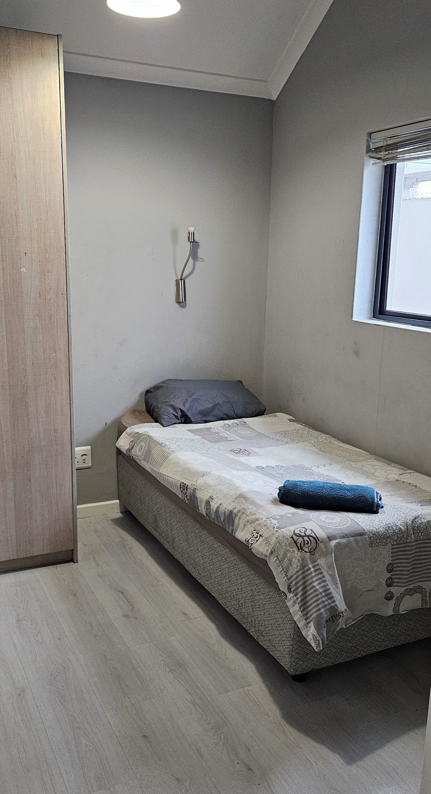 A small, single-bed room with a wooden door, a wall-mounted light fixture, and a window with blinds.