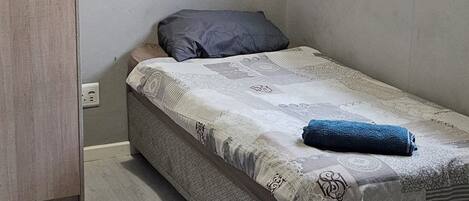 A small, single-bed room with a wooden door, a wall-mounted light fixture, and a window with blinds.
