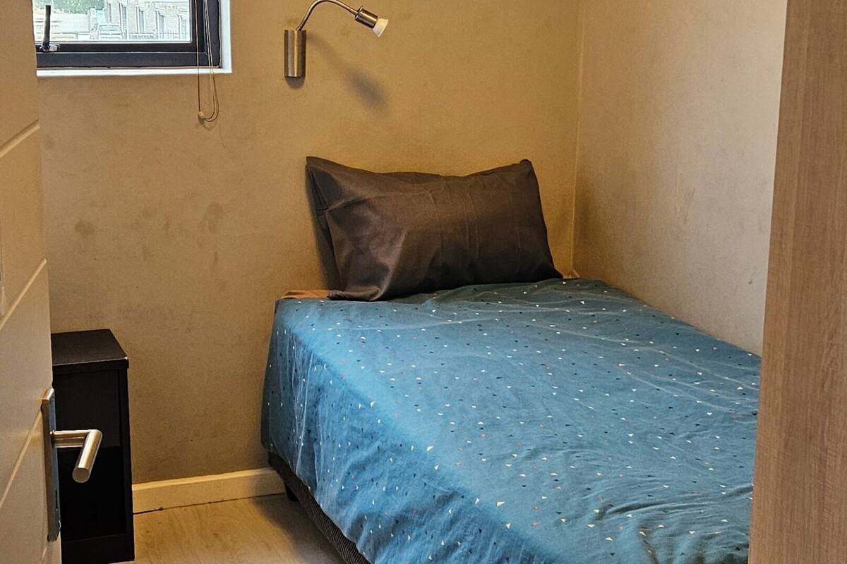 A small, single-bed room with a window, a bedside lamp, and a wooden floor.