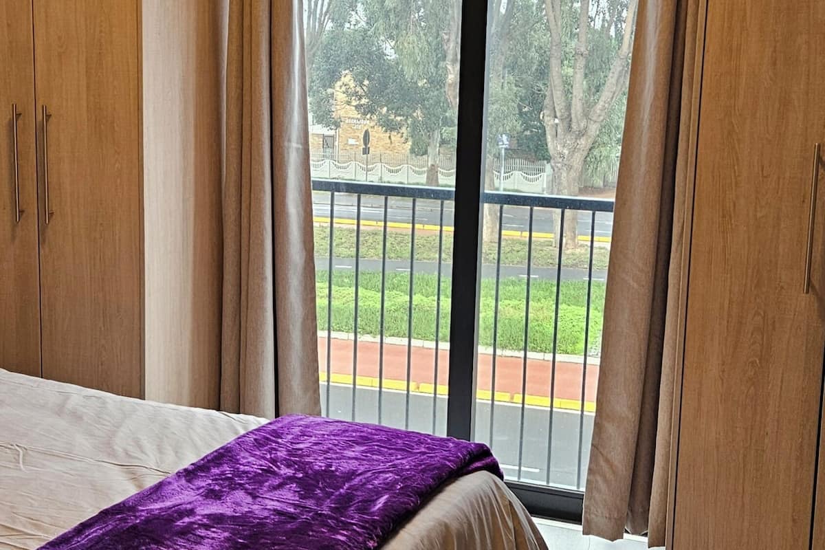 A bedroom with a large bed, a window with a view of a park, and wooden wardrobes.