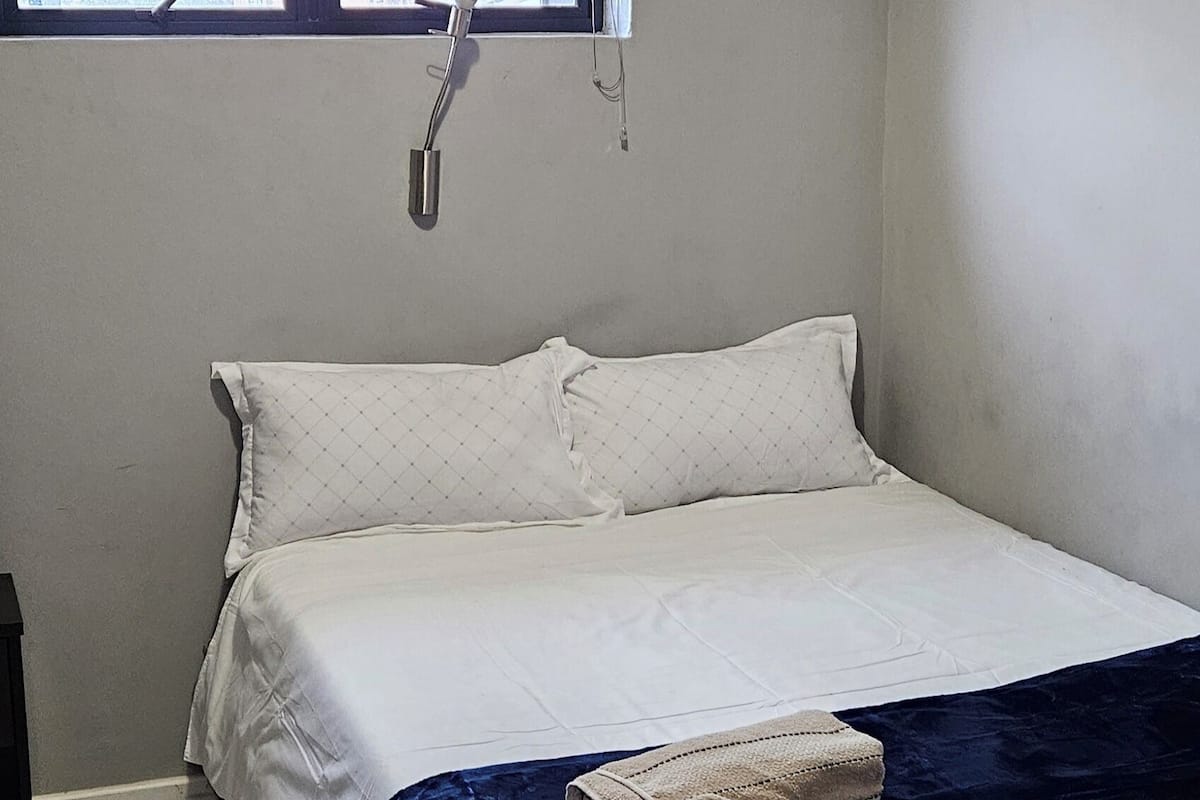 A single bed with a dark blue blanket, a white pillow, and a small towel on the footboard. A window with a metal handle is above the bed.