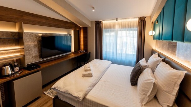 A modern hotel room with a large bed, a flat-screen TV mounted on the wall, a desk with a kettle, and a window with curtains. - Villa Bijoux (Ploiesti)