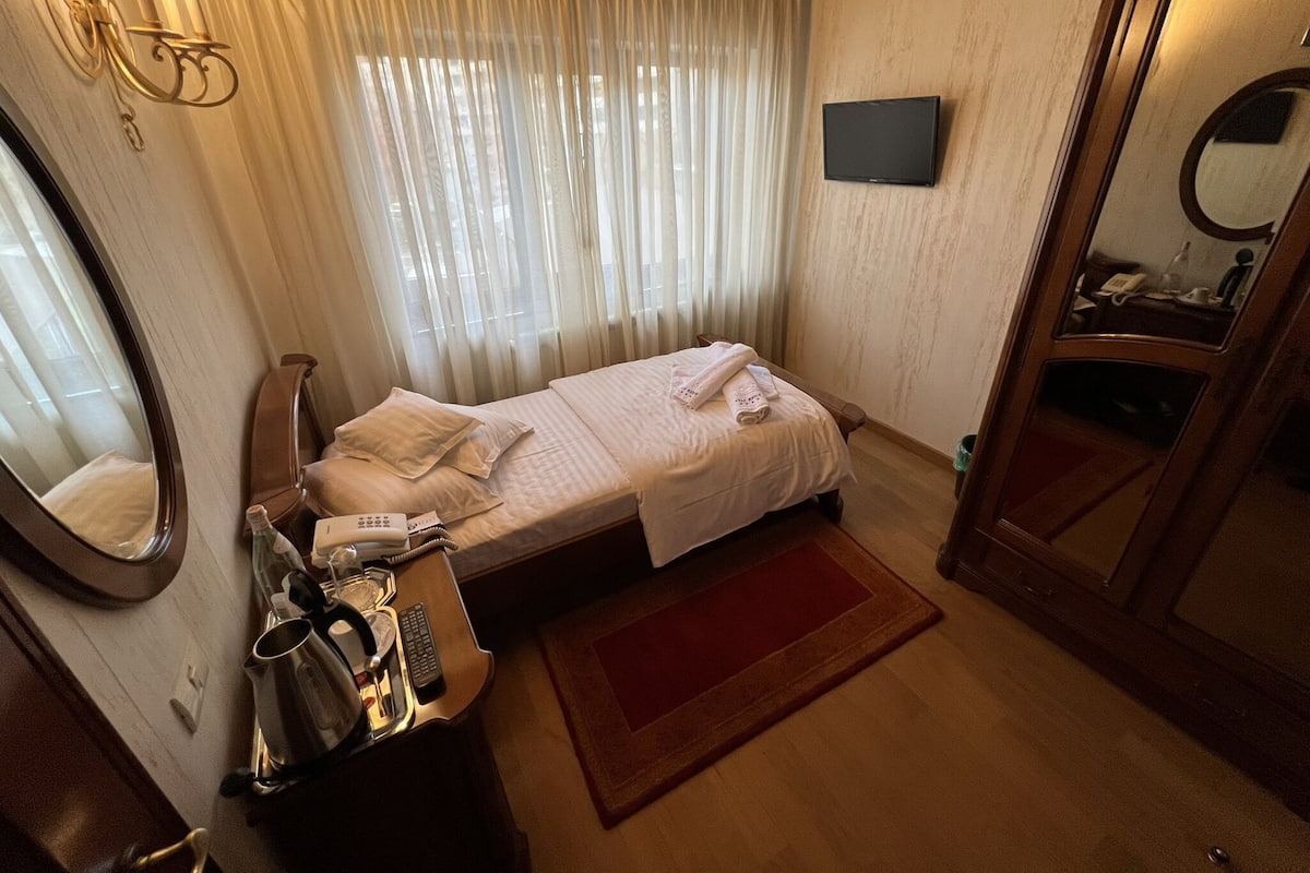 A hotel room with a single bed, a bedside table with a lamp, a mirror, a TV mounted on the wall, and a red rug.