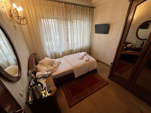 A hotel room with a single bed, a bedside table with a lamp, a mirror, a TV mounted on the wall, and a red rug. - Villa Bijoux (Ploiesti)