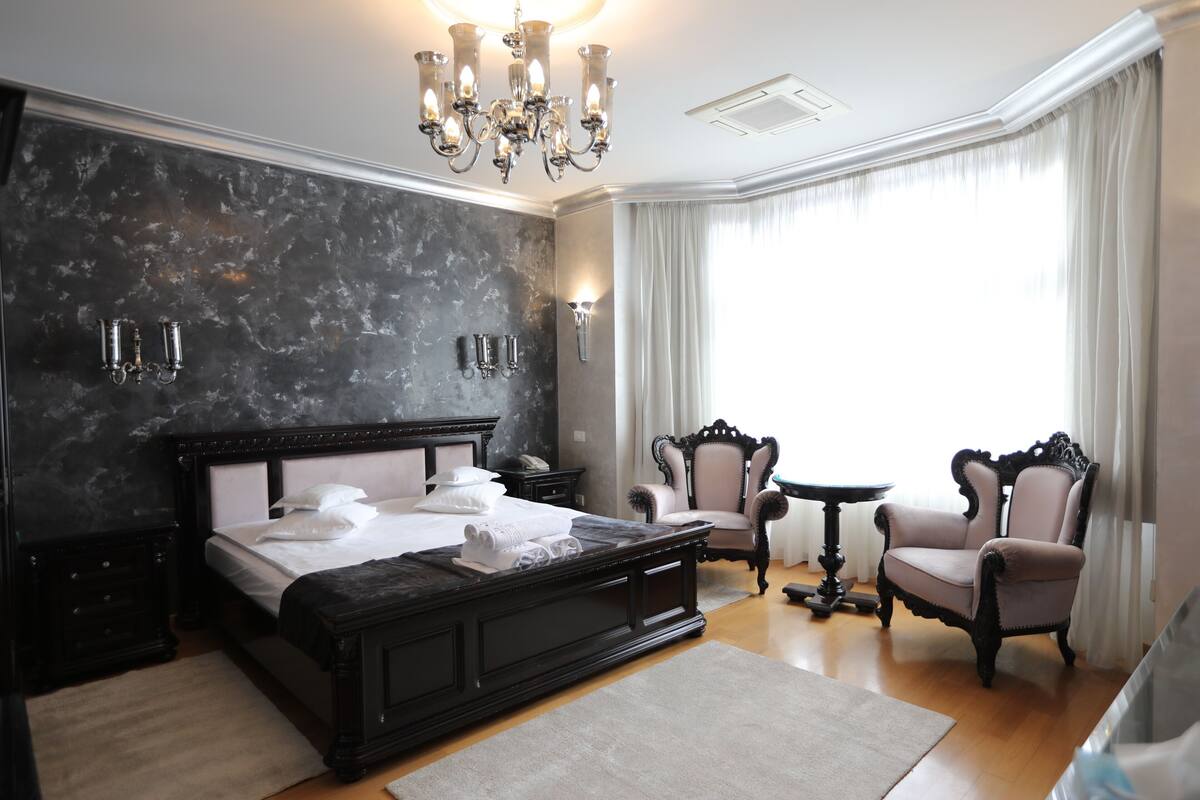 A spacious bedroom with a large bed, a chandelier, two armchairs, a small table, and a view of the outdoors through a window.