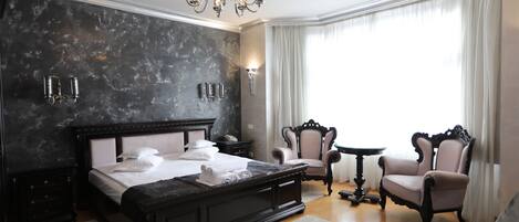 Un dormitorio amplio con una cama grande, una araña, dos sillones, una mesita y vistas al exterior a través de una ventana.