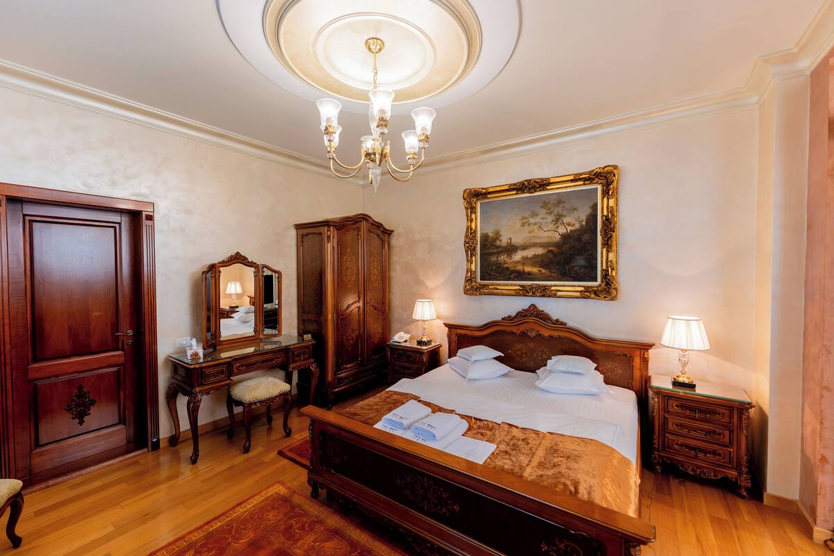 A hotel room with a large bed, wooden furniture, a chandelier, and a painting on the wall.