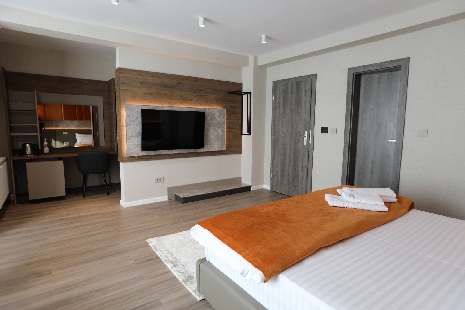A modern hotel room with a large bed, a TV mounted on a wooden wall, a desk with a chair, and a wardrobe.