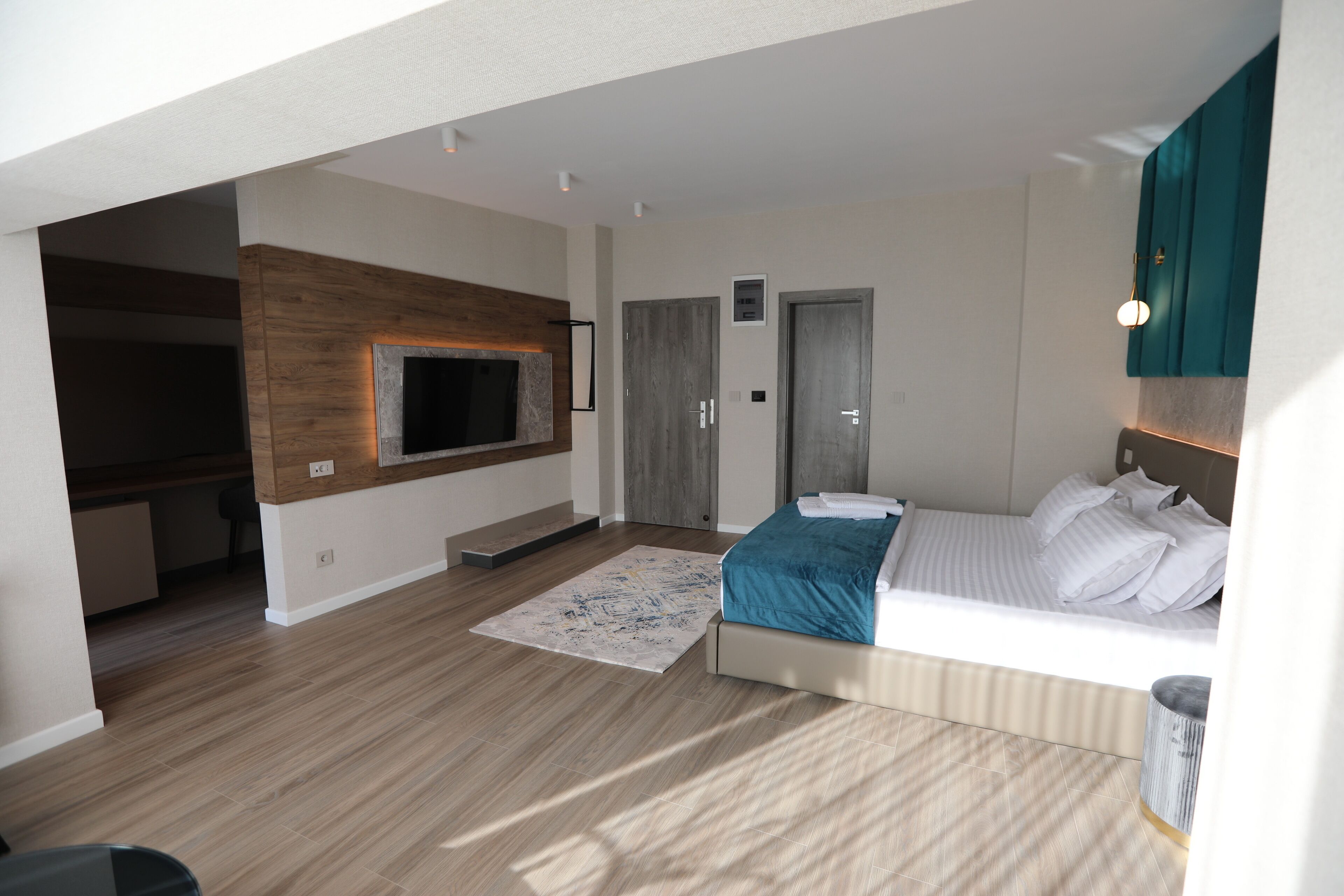 Ein modernes Schlafzimmer mit Bett, einem an einer Holzplatte montierten Fernseher, einem kleinen Teppich und einer Tür zu einem weiteren Raum.