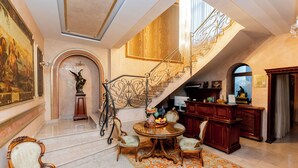A grand foyer with a marble floor, a staircase with ornate metal railings, a round wooden table with chairs, and a large painting on the wall. - Villa Bijoux (Ploiesti)