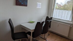 A small dining area with a white table and four black chairs. There is a window with a view of trees and buildings outside. - NestOaseWHV Küstenzimmer im Stadtherz (Wilhelmshaven)