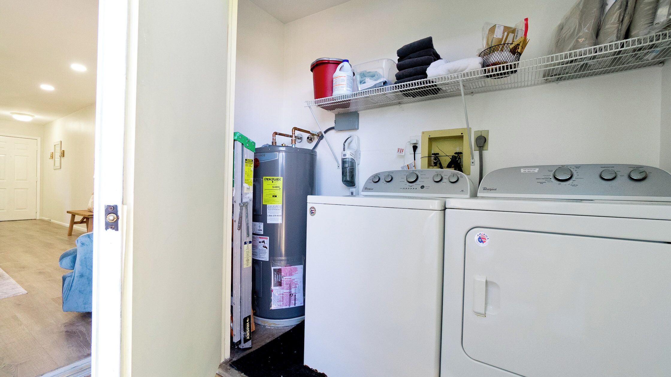 A compact laundry area with a washer and dryer, a water heater, and a shelf with towels and cleaning supplies.