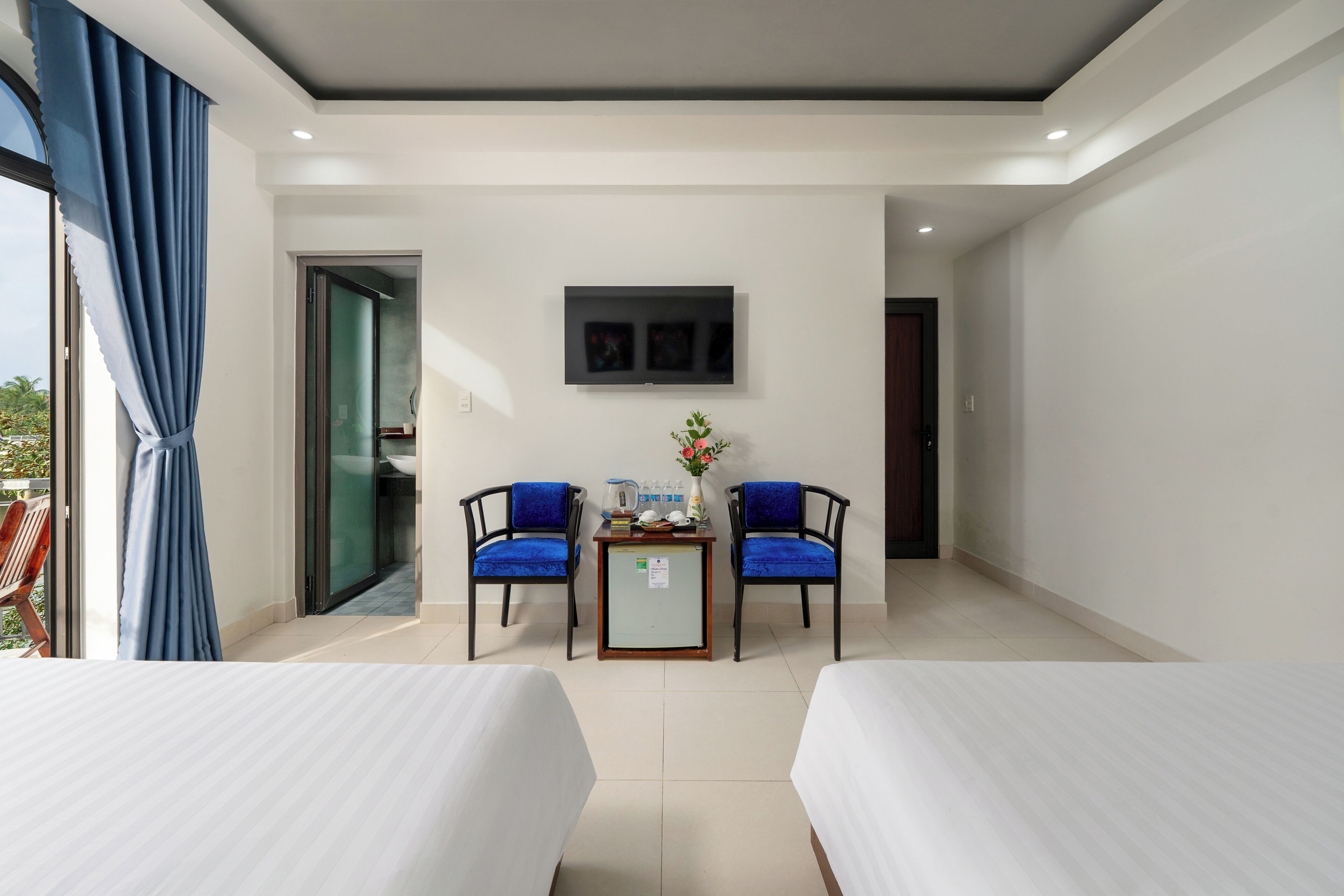 A hotel room with two beds, a small table with a vase, two blue chairs, and a TV on the wall.