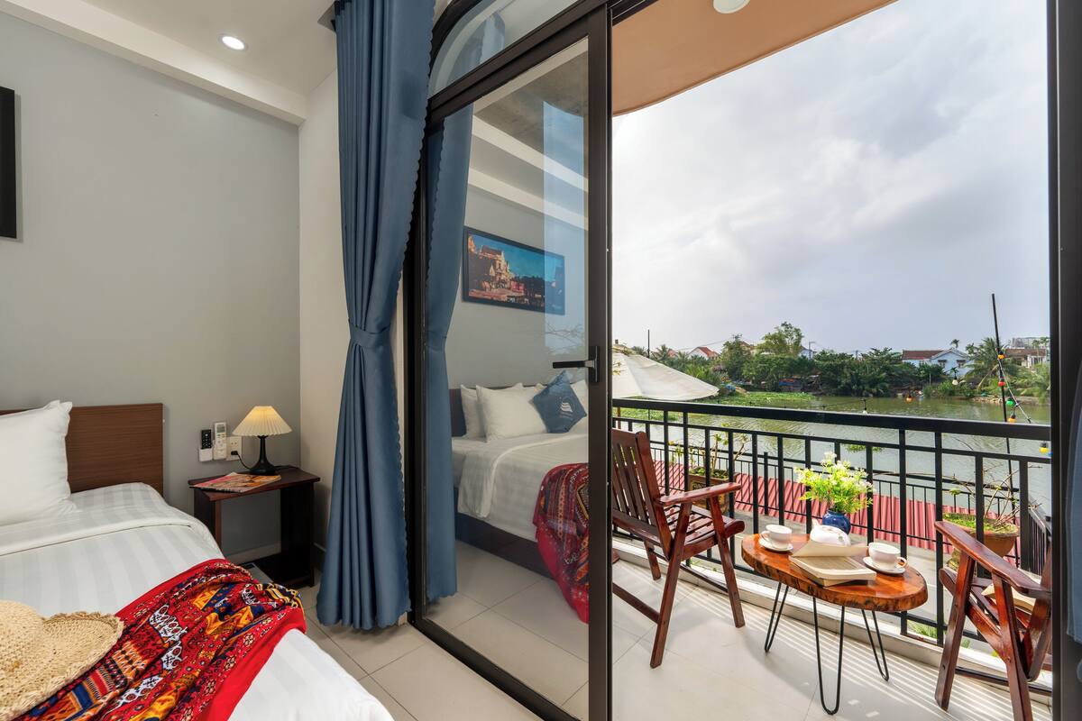 A hotel room with a bed, bedside table, lamp, and a balcony with a table, chairs, and a view of a river.