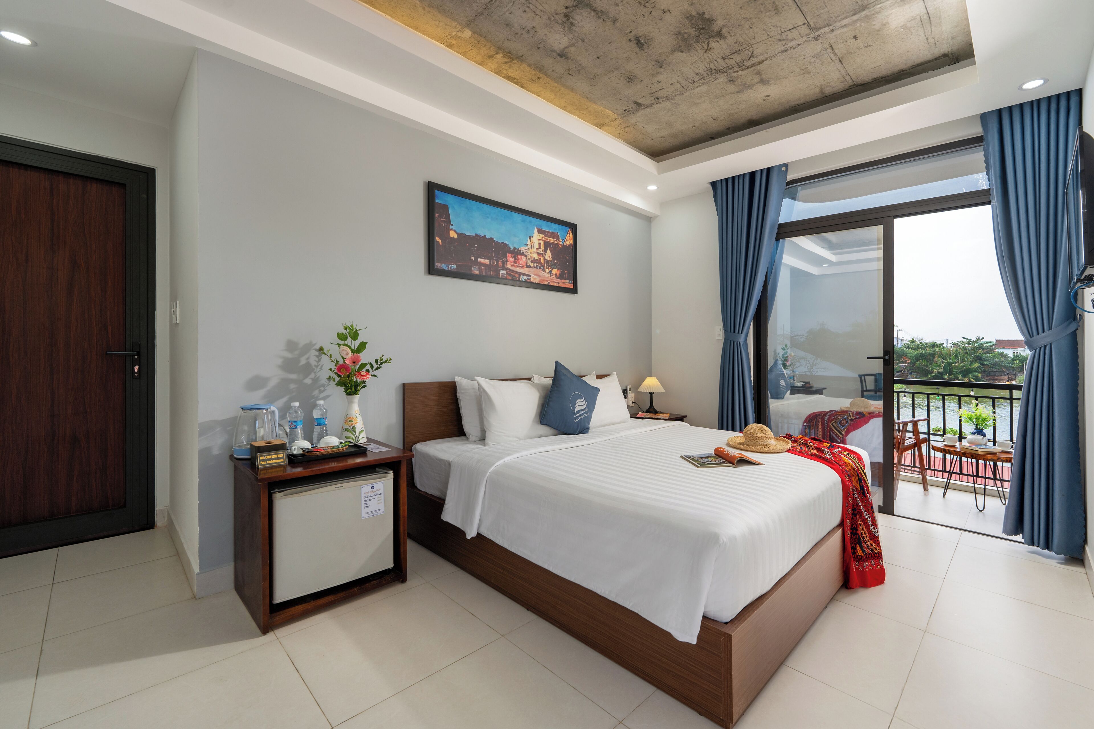 A hotel room with a bed, a nightstand, a desk, a chair, a balcony with a table and chairs, and a cityscape painting on the wall.