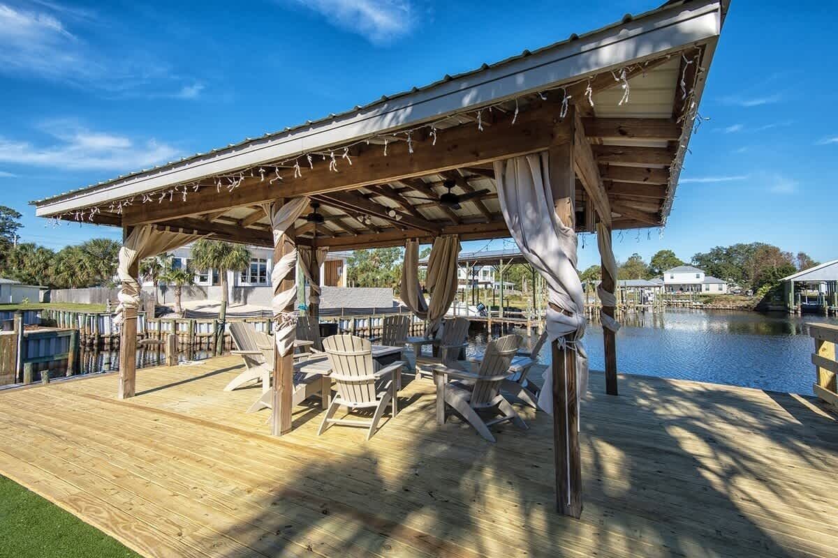 A wooden deck with Adirondack chairs, a pergola with string lights, and a view of a body of water.