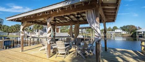 A wooden deck with Adirondack chairs, a pergola with string lights, and a view of a body of water.