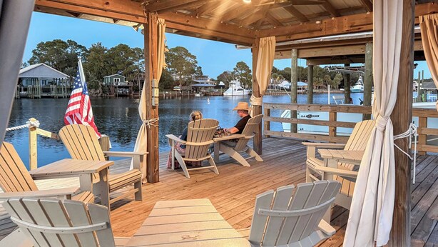 Een houten terras met Adirondack-stoelen en een tafel, met uitzicht op kalm water met boten en huizen op de achtergrond.