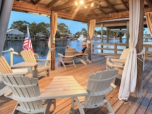 A wooden deck with Adirondack chairs and a table, overlooking a calm body of water with boats and houses in the background.