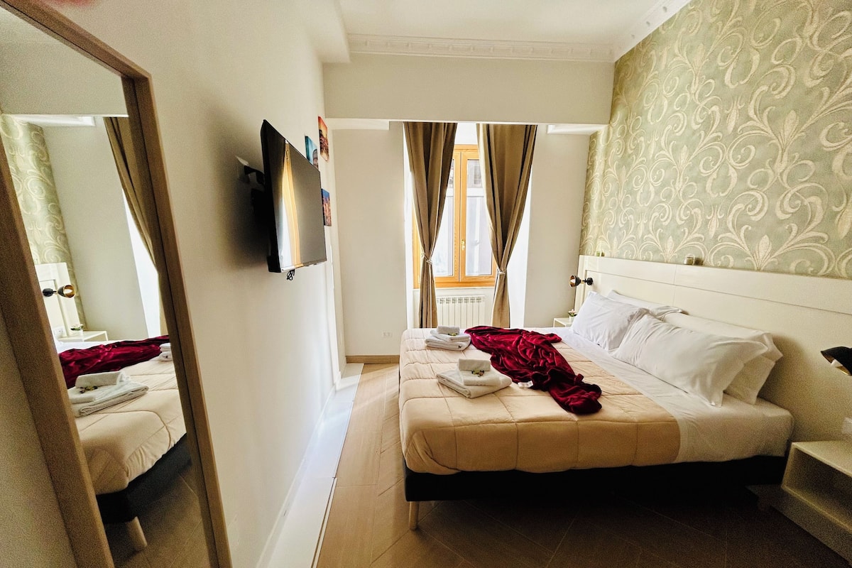 Een hotelkamer met een bed, een aan de muur bevestigde televisie, een raam met gordijnen en een spiegel die een deel van de kamer weerspiegelt.