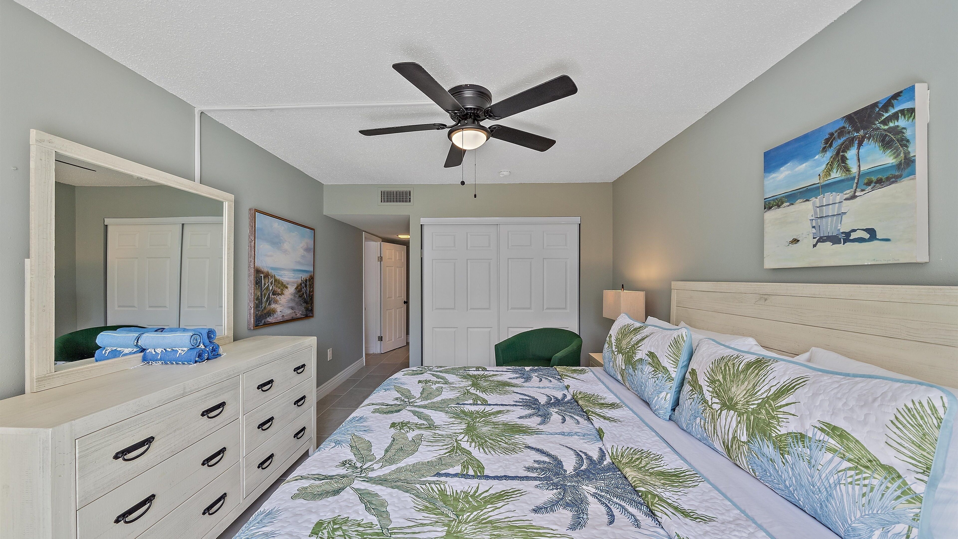 A bedroom with a bed, a ceiling fan, a dresser, a mirror, a door, and a painting.