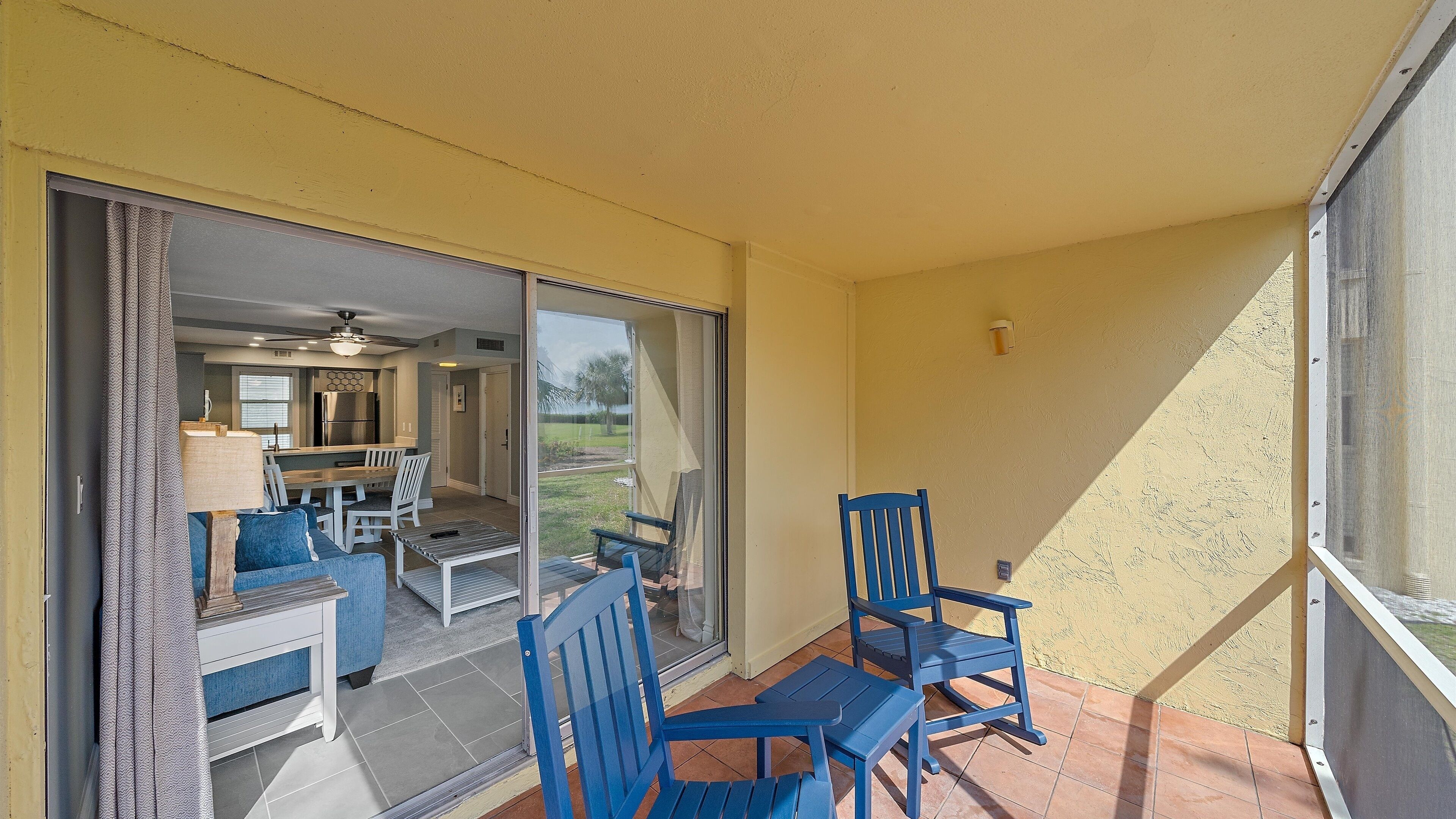 A patio with blue chairs and a table, a sliding glass door leading to a kitchen and dining area, and a yellow exterior wall.