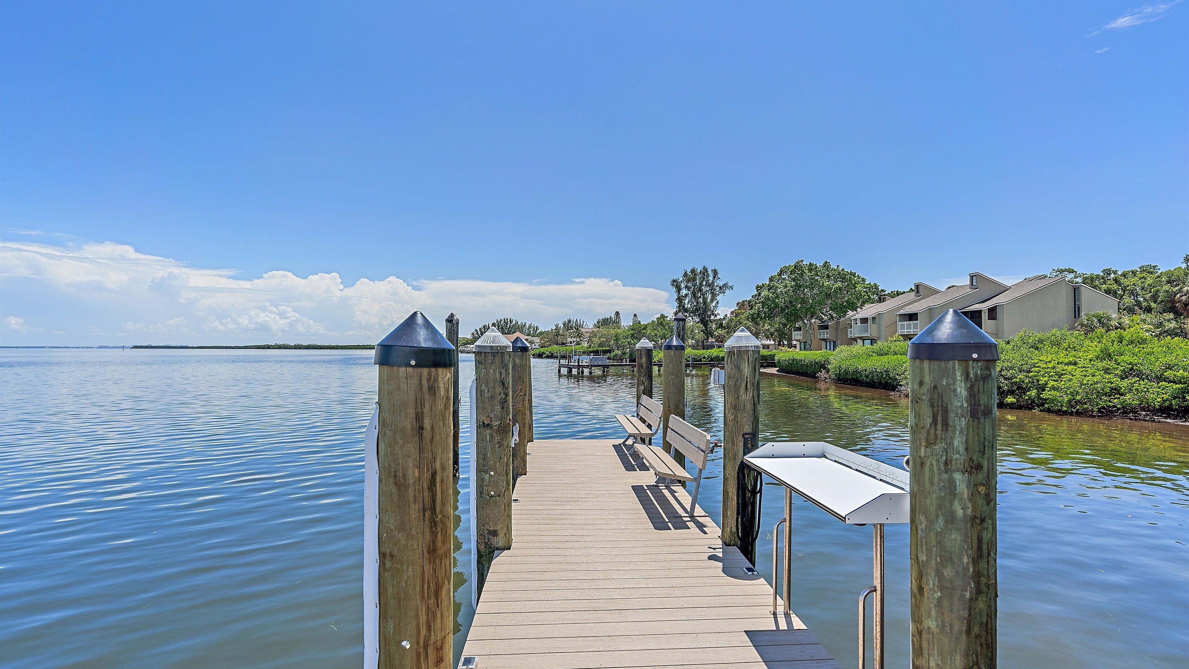 A wooden dock extending into calm water, with benches and a white sign, surrounded by greenery and residential buildings.