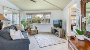 A living room with a grey sofa, a wooden armchair, a TV stand, and a large window with a view of the outside. - Kauai Plantation At Princeville 1121 3 Bedroom Condo (Princeville)